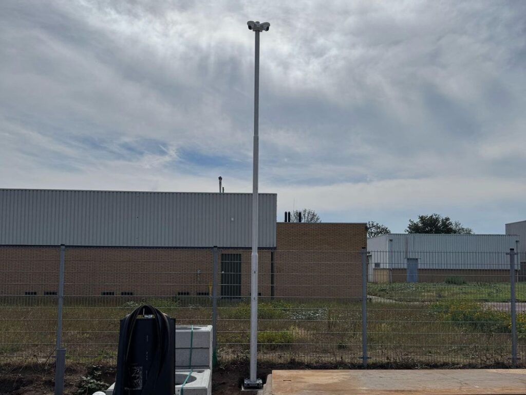 Hoge cameramast met twee camera's op een bouwterrein, over de volle hoogte in beeld, met hekwerk en bedrijfspanden erachter