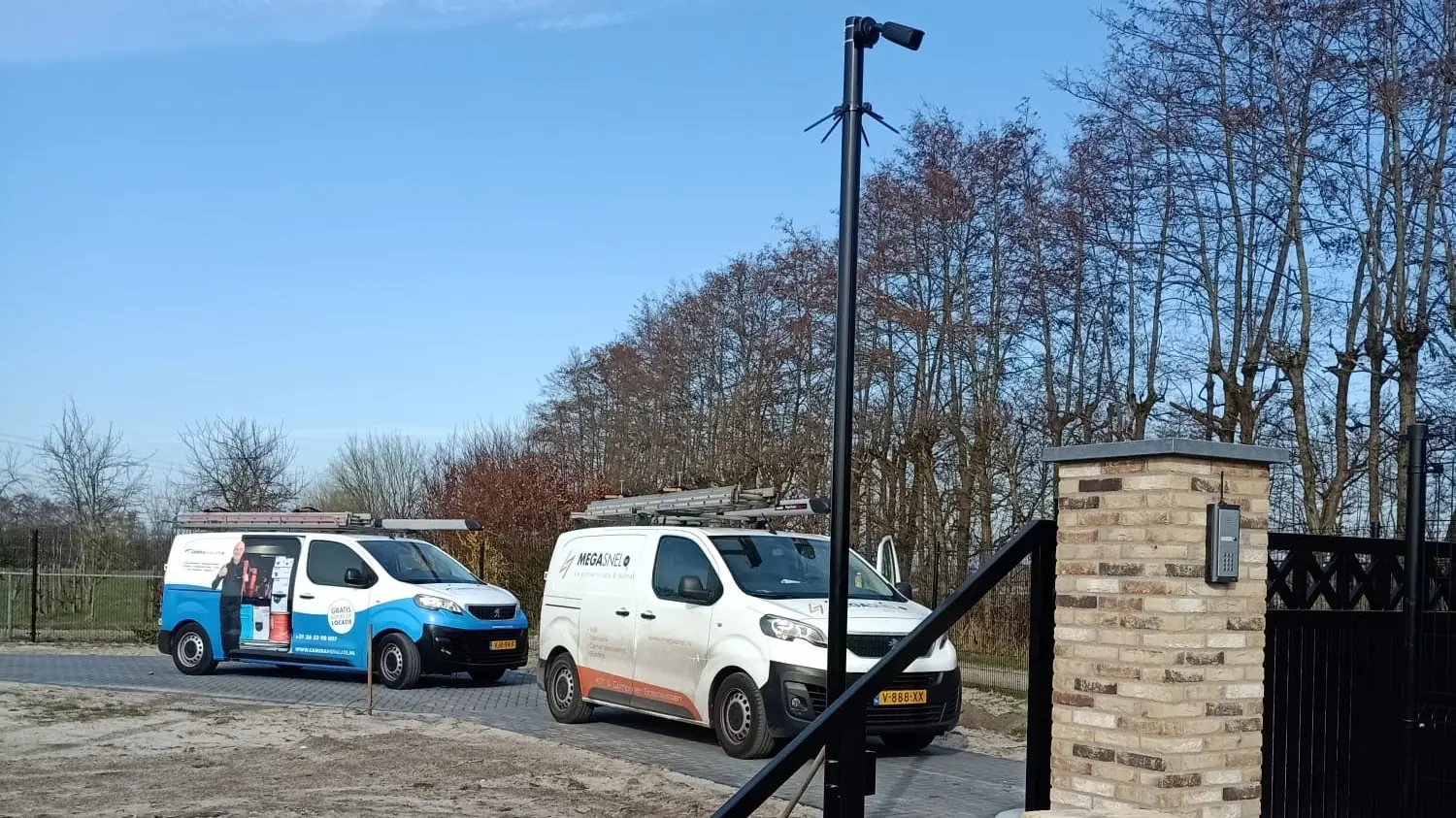 Camerapaal bij de oprit van een woning, met de bedrijfsbussen ernaast