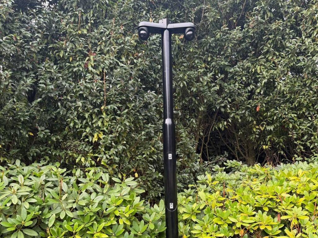 Zwarte ronde camerapaal met een dwarsarm en twee camera's, staand tussen hoge beplanting in een tuin