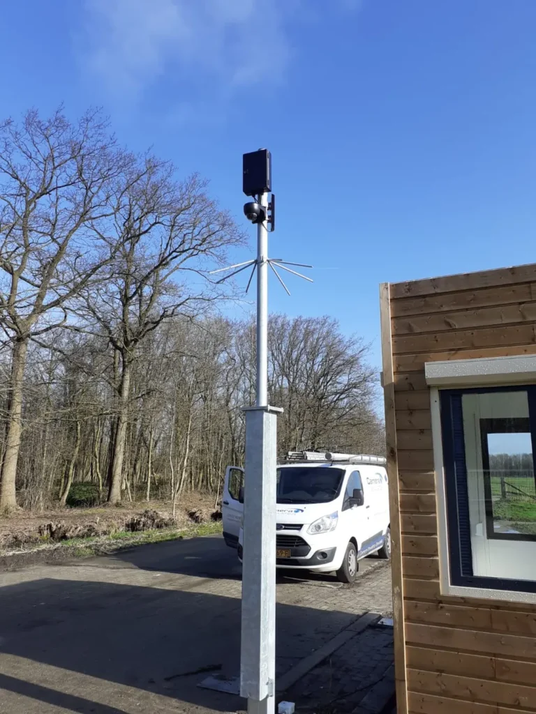 Lichtgrijze camerapaal naast een houten bedrijfspand, met de bedrijfsbus ernaast