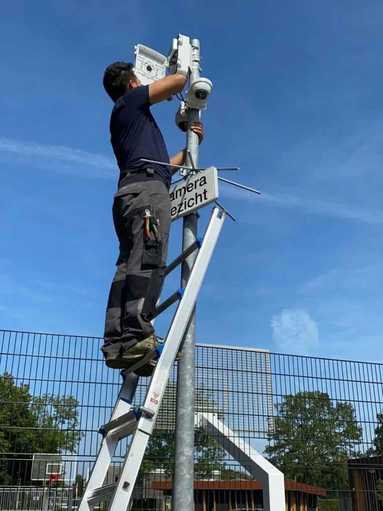 Monteur op een ladder aan de camerapaal langs een hekwerk, onder een blauwe lucht